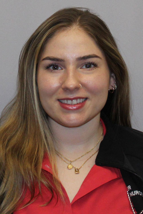 A woman with blonde hair is smiling for the camera, wearing a red outfit and a necklace. The background is a wall, and she has visible lipstick on her lips. Her layered hair frames her face, highlighting her features such as her eyebrows and chin.